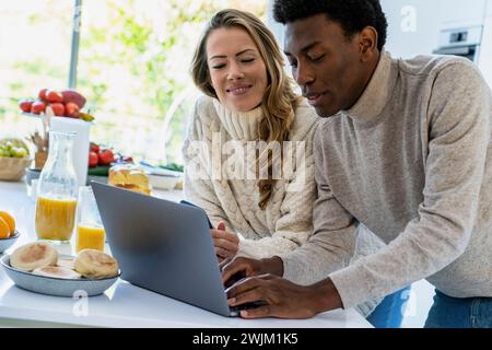 Adult man typing on laptop while leaning on kitchen counter with girlfriend Stock Photo