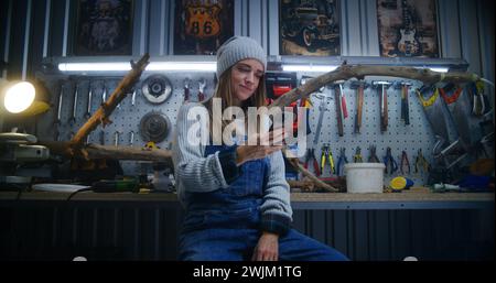 Female carpenter, craftswoman uses mobile phone sitting in stylish workshop. Wood product made from tree branch and board with professional tools in the background. Handicraft and entrepreneurship. Stock Photo