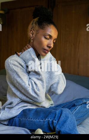 Stressed young adult woman touching her neck while sitting on bed Stock Photo