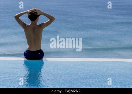 Young man sitting on edge of swimming pool near the sea Stock Photo