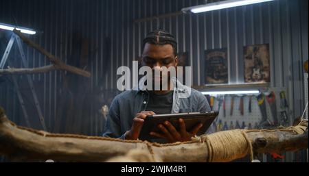 Creative African American designer uses digital tablet computer while creating design wooden products in modern garage. Craftsman or carpenter works in stylish workshop. Handmade and entrepreneurship. Stock Photo