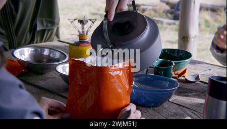 Close up of Caucasian woman taking kettle from gas burner and cooking freeze dried food in pouch. Group of tourists rests in wooden gazebo after walking trek. Hikers during expedition to mountains. Stock Photo