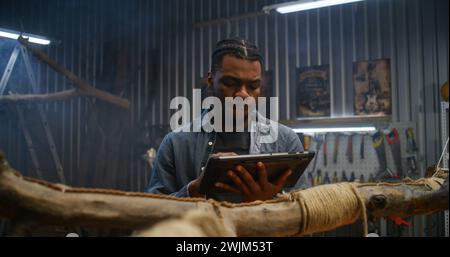 Creative African American designer uses digital tablet computer while creating design wooden products in modern garage. Craftsman or carpenter works in stylish workshop. Handmade and entrepreneurship. Stock Photo