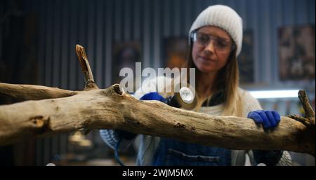 Female carpenter in goggles grinds tree branch using grinding machine. Craftswoman, designer wood processing, makes products from wood working in workshop. Hand crafted and small business concept. Stock Photo