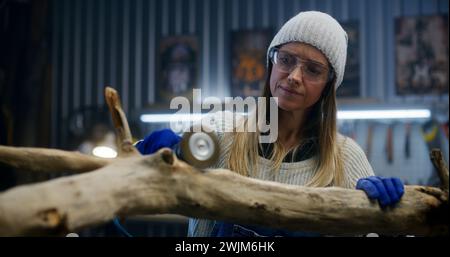 Female carpenter in goggles grinds tree branch using grinding machine. Craftswoman, designer wood processing, makes products from wood working in workshop. Hand crafted and small business concept. Stock Photo