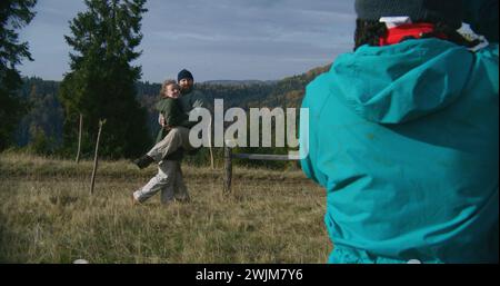 Young hiker takes pictures of Caucasian tourist family using camera. Happy couple of travelers posing on top of hill against beautiful landscape during hiking trip in mountains. Outdoor enthusiasts. Stock Photo