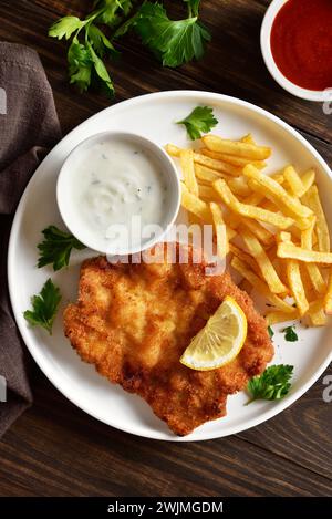 Homemade breaded wiener schnitzel served with potato fries and sauce on white plate over wooden background. Top view, flat lay Stock Photo