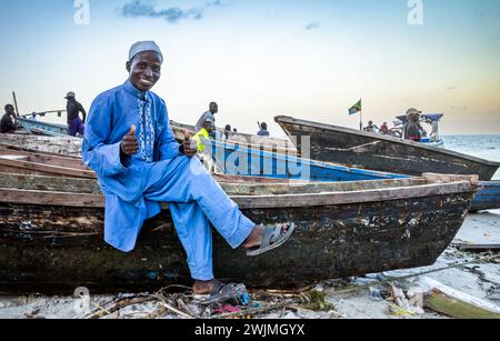 A man wearing a blue kanzu and white embroidered kofia smiles as he sits on a wooden dhow fishing boat at Kivukoni Fish Market in Dar es Salaam, Tanza Stock Photo