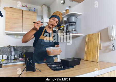 young latin man cook in uniform, at home making cake, he is happy with mixture he made with tutorial video on internet on phone, he is using mixer and Stock Photo