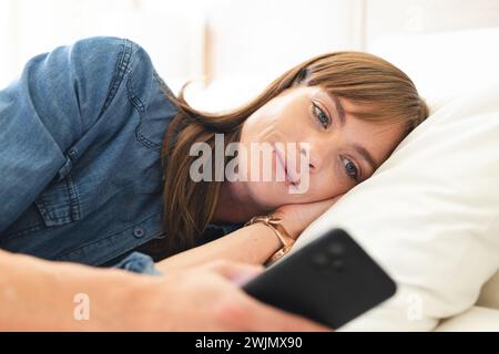 A middle-aged woman lies on a bed, gazing at her phone, appearing thoughtful Stock Photo