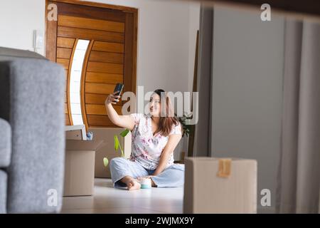 Young plus size biracial woman takes a selfie at home, surrounded by moving boxes, with copy space Stock Photo