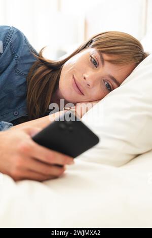 Woman in bed ponders, hinting at relaxation or digital engagement. Stock Photo