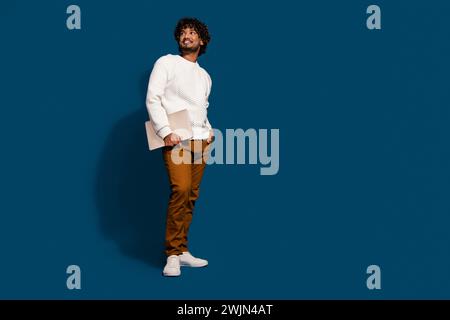 Full length photo of cool thoughtful guy dressed white pullover holding modern device looking empty space isolated dark blue color background Stock Photo