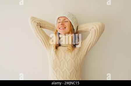 Relaxed enjoying happy smiling woman with closed eyes holds her hands behind her head in winter warm knitted clothes, sweater and hat Stock Photo