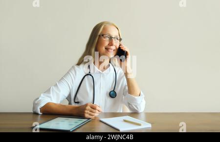 Caucasian middle-aged woman doctor calling on mobile phone with paper documents and digital tablet computer while sitting at desk in clinic office Stock Photo