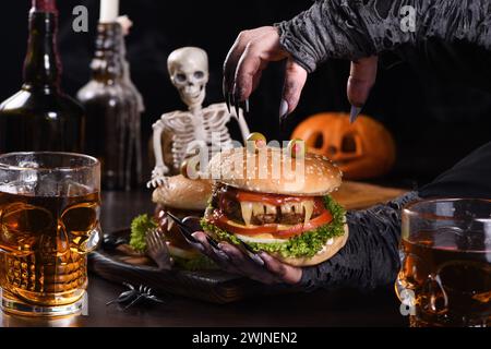 The witches hands hold a Monster Burger in her palms on a sitting skeleton. Perfect Halloween Party appetizer Stock Photo