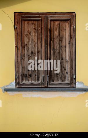 Yellow painted house wall with cracked surface, two windows. Window with brown shutters on a yellow wall. Stock Photo