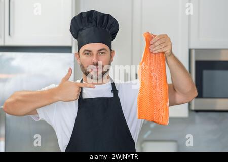 Portrait of chef man hold fish salmon in a chef cap in the kitchen. Man wearing apron and chefs uniform and chefs hat. Raw fish salmon fillet for adve Stock Photo
