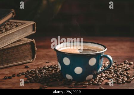 Steaming blue coffee cup surrounded by coffee beans and old books on a wooden table Stock Photo