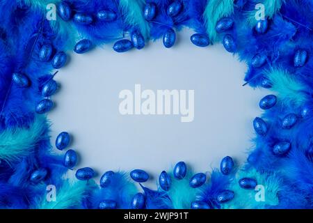 Frame made of blue and turquoise feathers with blue chocolate eggs on a light blue background Stock Photo