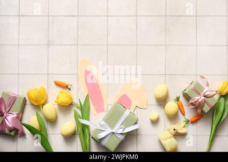 Paper bunny ears with Easter eggs, tulip flowers and gift boxes on white tiled background Stock Photo