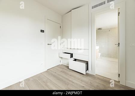Corner of a newly renovated empty bedroom with white painted walls and a built-in wardrobe with white wooden doors and drawers next to an en-suite bat Stock Photo