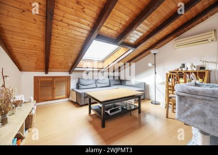 A small living room with wooden sloping ceilings with a large integrated skylight, a wooden desk and Venetian-style wooden doors in the lower part of Stock Photo
