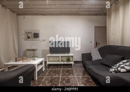 Small living room with white furniture, black fabric sofa, TV on a sideboard and dark stoneware on the floor Stock Photo