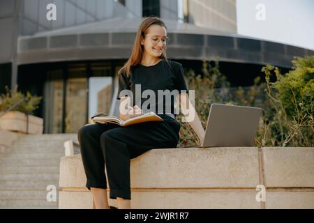 Pretty businesswoman making notes and browsing laptop outdoors near city building Stock Photo