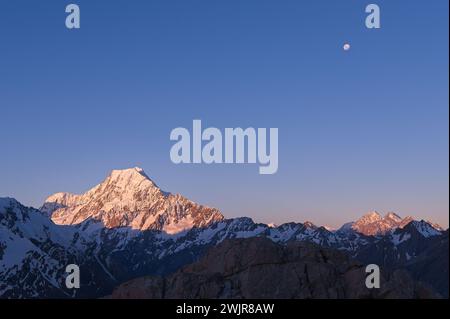 Scenic Sunrise at Mount Cook: Snowy Mountain Peaks Glowing in Golden Light, Accompanied by the Moon in New Zealand's Breathtaking Landscape Stock Photo