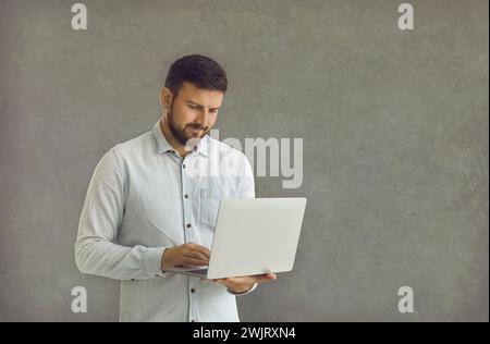 Young and confident man standing on a gray background with a laptop in his hands. Stock Photo