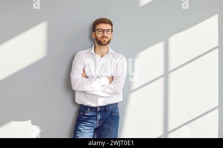 Confident bearded man in eyeglasses posing with crossed arms at gray wall Stock Photo