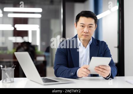 Focused professional businessman in a suit using a tablet at his desk with a laptop in a contemporary office setting. Stock Photo