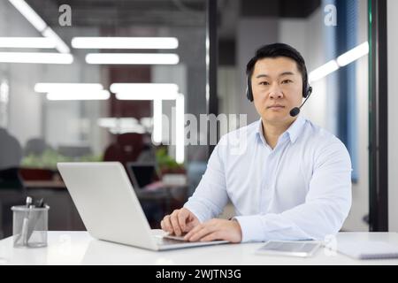 Focused Asian man in smart casual wear using laptop at desk with headset in a contemporary office setting. Stock Photo