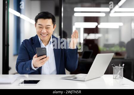 Happy Asian executive engaging in a virtual greeting, waving at the camera while having a video call on his mobile phone. Stock Photo