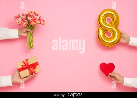 Man hands with balloon in shape of figure 8, bouquet of tulips, paper heart and gift box on pink background. International Women's Day celebration Stock Photo