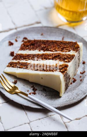 A piece of carrot cake served on white tile table. bright sunny scene. Layered cake with cream cheese frosting and fluffy biscuit with drink Stock Photo