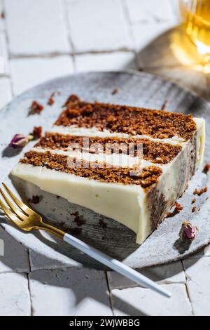 A piece of carrot cake served on white tile table. bright sunny scene. Layered cake with cream cheese frosting and fluffy biscuit with drink Stock Photo