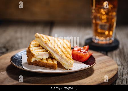Sliced Cut Toast sandwich with molten Gouda cheese and turkey ham on a wooden background with cherry tomato on a black plate. Served with ice tea. Stock Photo