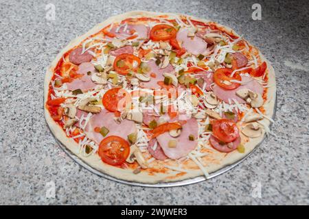 Uncooked pizza with sausage, ham, mushroom, pepper, tomatoes mozzarella on table in pizzeria or home. Italian cuisine, homemade bakery, fast food, sem Stock Photo