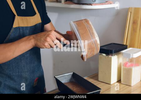 close-up unrecognizable dark-haired man in home kitchen standing with hand mixer pouring cake mixture into baking pan, pastry concept, copy space. Stock Photo