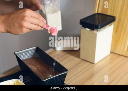 unrecognizable young man's hand at kitchen table is placing sugar with plastic spoon into cake mixture inside baking pan Stock Photo