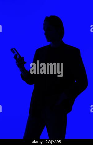 Silhouette an elegant businessman using a modern smartphone in a jacket standing on a blue glowing background, a futuristic concept Stock Photo