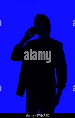 Unrecognizable Silhouette an elegant businessman talking on a smartphone in a jacket standing on a blue glowing background, a futuristic concept Stock Photo