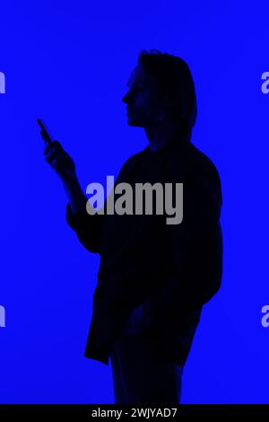 Silhouette an elegant businessman using a modern smartphone in a jacket standing on a blue glowing background, a futuristic concept Stock Photo