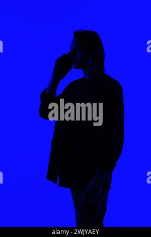 Unrecognizable Silhouette an elegant businessman talking on a smartphone in a jacket standing on a blue glowing background, a futuristic concept Stock Photo