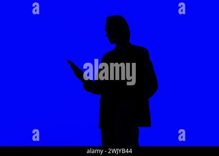 Silhouette an elegant businessman using a modern smartphone in a jacket standing on a blue glowing background, a futuristic concept Stock Photo