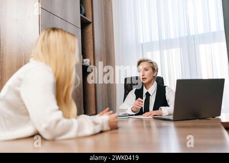 View from table to professional middle-aged female teacher, manager or mentor helping young blonde student, new employee, teaching intern, explaining Stock Photo