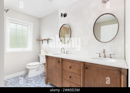 A bathroom with a brown tiled wall with lights mounted over circular mirrors, natural wood cabinet, and a blue and white mosaic tile floor. Stock Photo