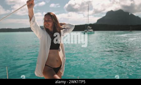 Happy female relax on yacht tour enjoy seascape in sunny day. Young woman in bikini stands on boat nose, turquoise water and tropical green island. Travel summer holiday vacation. French Polynesia Stock Photo